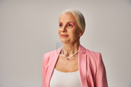 femme âgée rêveuse en veston rose et collier de perles souriant et regardant loin sur le gris, élégance