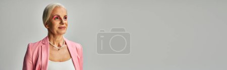 dreamy and stylish mature lady in pink blazer and pearl necklace looking away on grey, banner