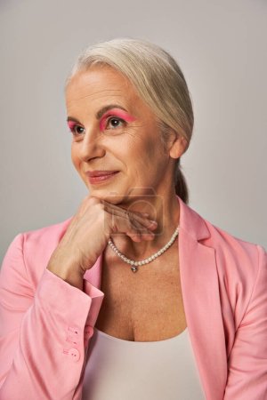 Photo for Happy and dreamy senior model in pink elegant blazer holding hand near chin and looking away on grey - Royalty Free Image