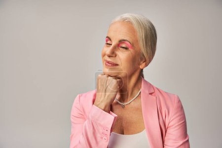 dreamy and smiling senior woman in pink blazer holding hand near and posing with closed eyes on grey
