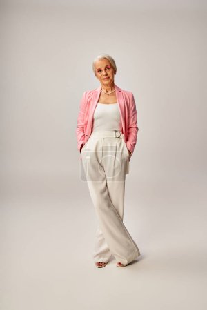 full length of mature lady in pink blazer posing with hands in pockets of white pants on grey