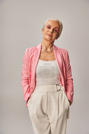 trendy mature lady in pink blazer standing with hands in pockets of white pants on grey, elegance