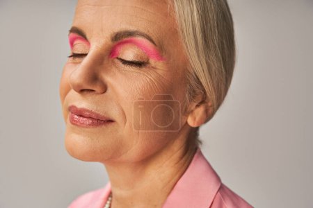Nahaufnahme Porträt einer stilvollen Seniorin mit natürlichem Make-up, die mit geschlossenen Augen auf grau lächelt