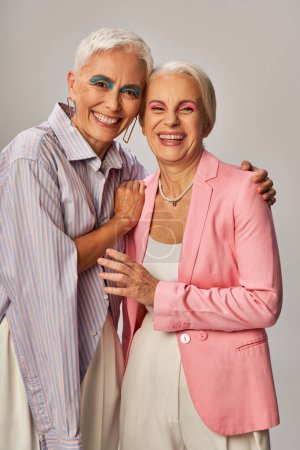 alegre senior mujeres en elegante azul y rosa ropa abrazando y sonriendo a la cámara en gris