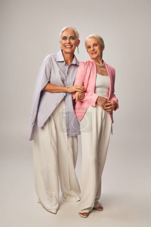 fashionable and joyful senior women in stylish casual attire smiling at camera on grey, full length