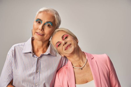 joyful senior female friends in trendy blue and pink attire looking at camera on grey, happiness