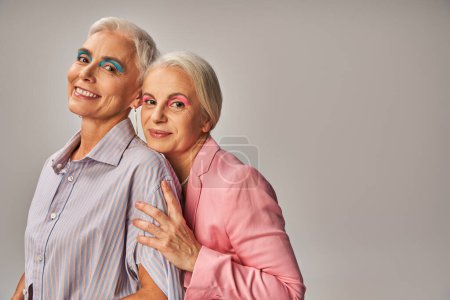 delighted senior female friends in blue and pink clothes smiling at camera on grey, age positivity