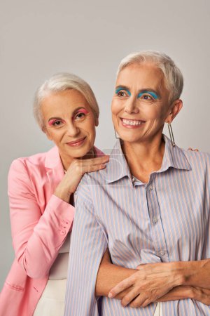 Glückliche Seniorin blickt in die Kamera neben trendiger Freundin mit blauem Eyeliner vor grauem Hintergrund