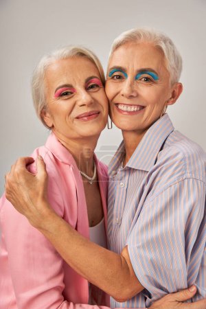 glückliche Seniorin mit blauem Eyeliner umarmt elegante Freundin auf grau, modisches Altern