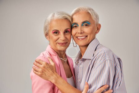 modische Seniorinnen in blauer und pinkfarbener Kleidung umarmen und lächeln in die Kamera auf grau