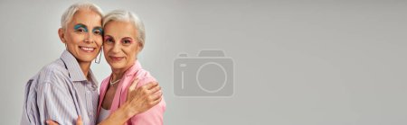 vanidad estilo justo, felices modelos senior en traje azul y rosa abrazando sobre fondo gris, bandera