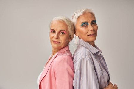 Glamour-Seniorinnen in stilvoller blauer und pinkfarbener Kleidung stehen Rücken an Rücken vor grauem Hintergrund