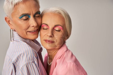 portrait of mature female friends with silver hair and makeup standing with closed eyes on grey