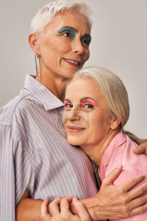 Trendige Seniorin mit blauem Eyeliner umarmt zufriedene Freundin und blickt in Kamera auf grau