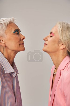 Foto de Elegantes amigas mayores en ropa azul y rosa de pie con los ojos cerrados en gris, vista lateral - Imagen libre de derechos