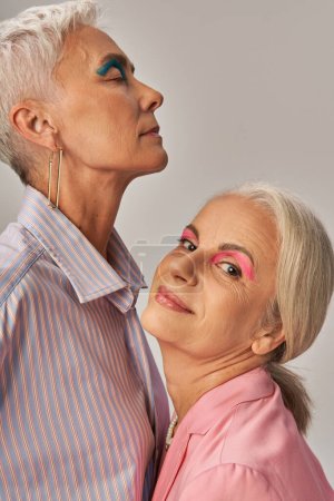 pleased senior woman looking at camera near trendy female friend with blue makeup on grey backdrop