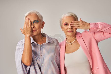 trendy mature models in blue and pink wear obscuring faces with hands and looking at camera on grey