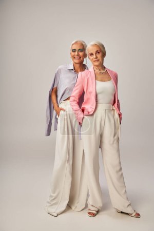 in voller Länge glückliche Seniorinnen in trendiger Kleidung, die Hände in Taschen auf grau