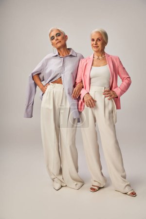 in voller Länge lächelnde Seniorinnen in stylischer Kleidung posieren mit den Händen auf den Hüften auf grau
