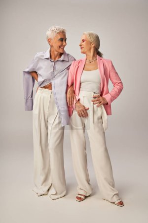 amis féminines seniors à la mode debout avec les mains sur les hanches et souriant à l'autre sur le gris