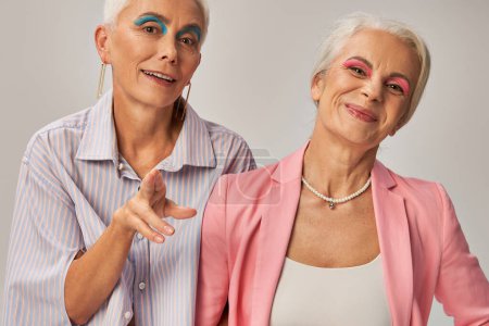 Foto de Mujer mayor de moda con delineador de ojos azul apuntando con los dedos a la cámara cerca de alegre amigo en gris - Imagen libre de derechos