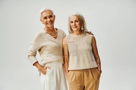 stylish aging, cheerful female friends in trendy casual attire posing with hands in pockets on grey