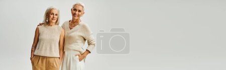 Téléchargez les photos : Joyeuses dames mûres en vêtements pastel à la mode debout avec les mains dans les poches sur gris, bannière - en image libre de droit
