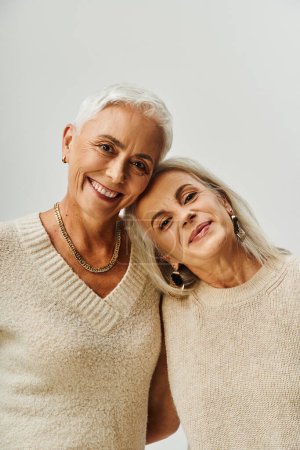 Porträt lächelnder Seniorinnen in Make-up und goldenen Accessoires, die vor grauem Hintergrund in die Kamera schauen