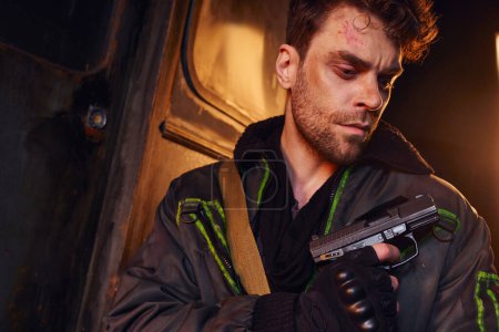 unshaven and gloomy man with scratched face looking at gun in dark subway, post-disaster refuge