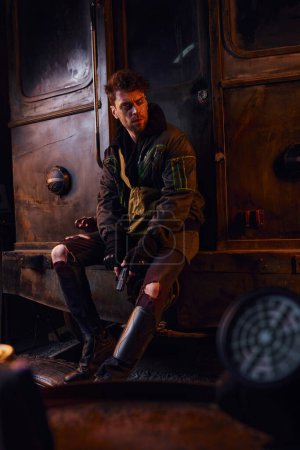 devastated man in worn clothes sitting with gun on rusty subway carriage, post-apocalyptic isolation