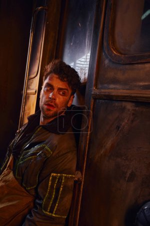 Unrasierter Mann in Jacke schaut in der Nähe rostiger Waggons in Gefahrenumgebung verlassener U-Bahn weg