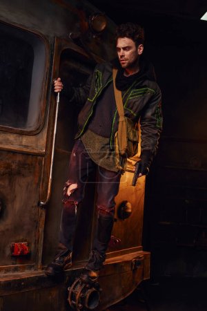 Mann in verschlissenem Outfit steht mit Waffe auf rostigem U-Bahn-Waggon, postapokalyptisches Leben, volle Länge