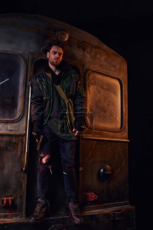 post-apocalyptic survivor in worn clothes standing with gun on rusty subway carriage in underground
