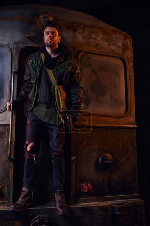 Mann in verschlissenem Outfit hält Waffe in der Nähe rostiger U-Bahn-Waggons und blickt in die Kamera, Leben nach der Katastrophe