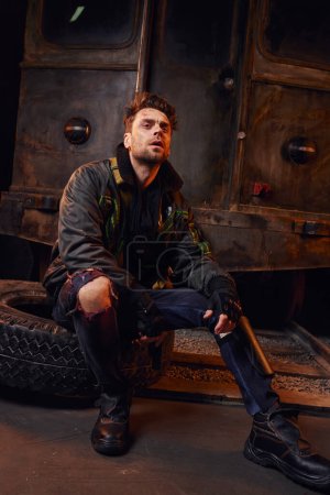 Unrasierter Mann in verschlissenem Outfit sitzt auf Reifen neben rostigem Wagen in U-Bahn nach Katastrophe, in voller Länge