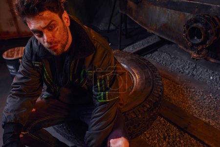 Téléchargez les photos : Homme désespéré avec visage rayé non rasé assis sur un pneu dans l'obscurité du métro post-apocalyptique - en image libre de droit
