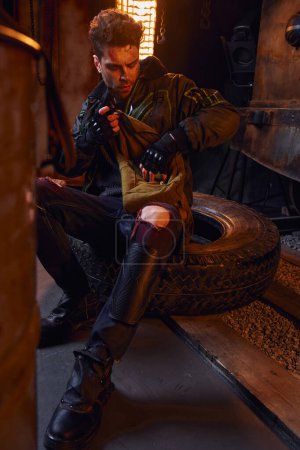 Téléchargez les photos : Homme en vêtements usés et gants sans doigts assis sur un pneu avec sac en toile dans le métro post-apocalyptique - en image libre de droit