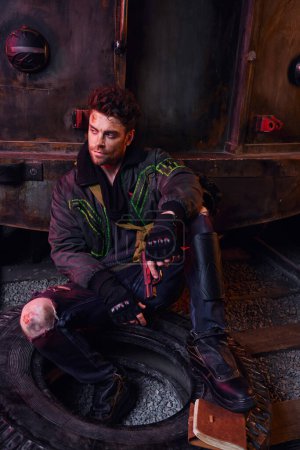 unshaven man with gun sitting on tire near diaries in dark subway, post-apocalyptic concept