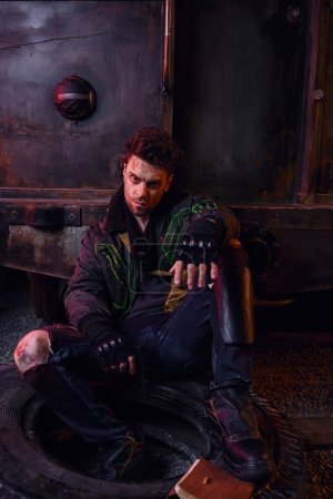 grimmiger Mann in verschlissenem Outfit sitzt auf Reifen mit Waffe in verlassener U-Bahn, Leben nach der Atomkatastrophe
