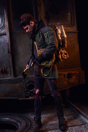 Foto de Hombre armado con arma y hacha de pie con mochila y botella de agua en oscuro abandonado subterráneo - Imagen libre de derechos