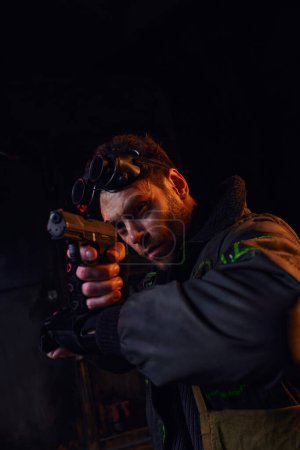 hombre con googles militares mirando hacia otro lado y apuntando con el arma en la luz roja de post-desastre subterráneo