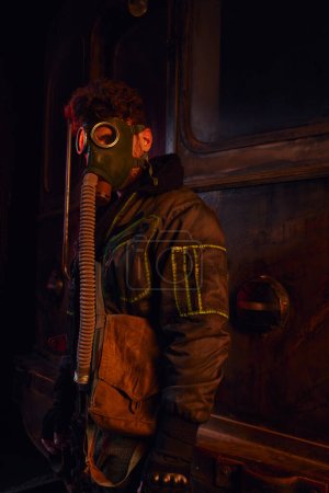 Mann in schmutziger Jacke und Gasmaske nahe rostigem U-Bahn-Waggon in verlassenem U-Bahn-Tunnel