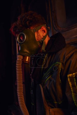 Foto de Hombre con máscara y chaqueta desgastada en luz roja de túnel subterráneo oscuro, supervivencia post-apocalíptica - Imagen libre de derechos