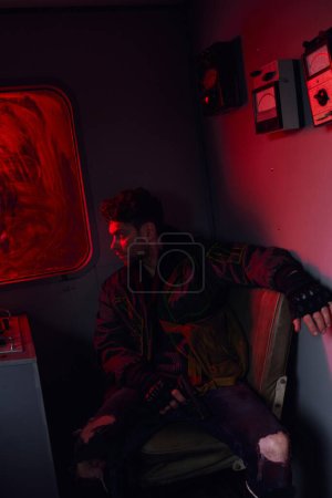 Téléchargez les photos : Homme désespéré avec arme dans un wagon de métro sale avec lumière rouge, personnage de jeu post-apocalypse - en image libre de droit