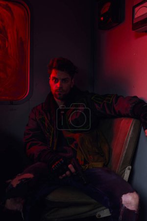 depressed man with gun sitting in abandoned subway carriage in red light, post-apocalyptic concept