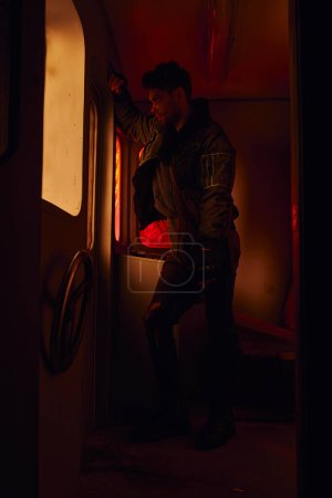 Foto de Hombre con arma mirando a través de la ventana del carruaje abandonado en el metro post-desastre con luz roja - Imagen libre de derechos