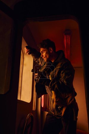 Foto de Hombre con pistola de pie cerca de la ventana de carro abandonado en el metro post-apocalíptico con luz roja - Imagen libre de derechos