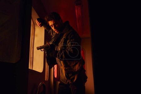 düsterer Mann mit Waffe blickt in verwüsteten U-Bahn-Waggon mit Rotlicht in die Kamera, Post-Apokalypse