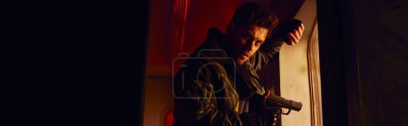 Photo for Unshaven man with gun looking at camera in abandoned subway carriage with red light, banner - Royalty Free Image