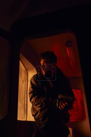 concentrated man with gun looking at camera in subway carriage in post-disaster subway in red light